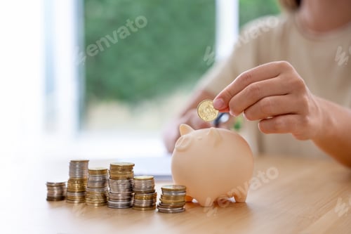 Preview: Woman putting coin into piggy bank at wooden table indoors, closeup