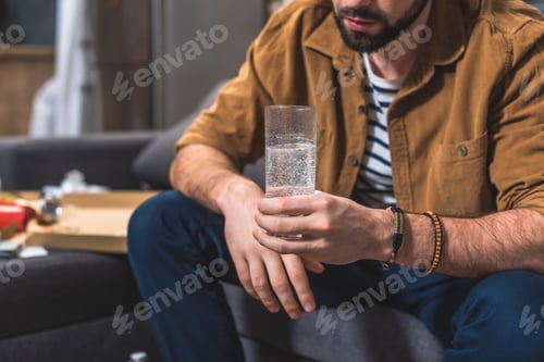 Preview: cropped image of loner with hangover holding glass of water at living room