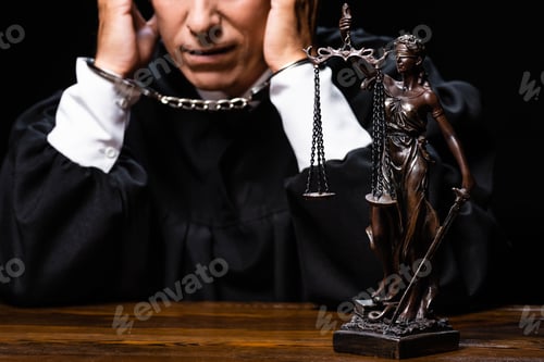 Preview: cropped view of judge in judicial robe sitting at table with handcuffs isolated on black