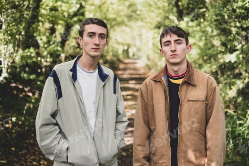 Preview: Young man and teenage boy with short brown hair wearing casual jackets standing side by side on a