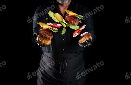 Preview: Chef in a black shirt, apron stands on a black background, flying cheeseburger ingredients