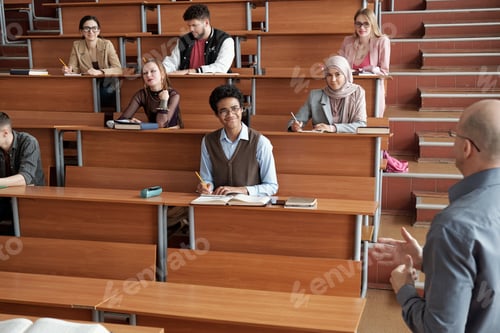 Preview: Happy intercultural university students making notes and listening to professor at lesson