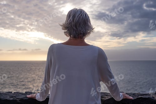 Preview: Rear view of senior woman standing in front of the sea at sunset looking the horizon over the water