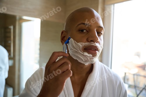 Preview: Serious young man shaving with razor and looking at camera in bathroom