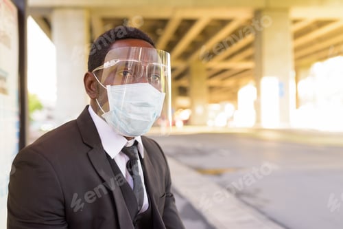 Preview: African businessman with mask and face shield waiting at the bus stop in the city outdoors