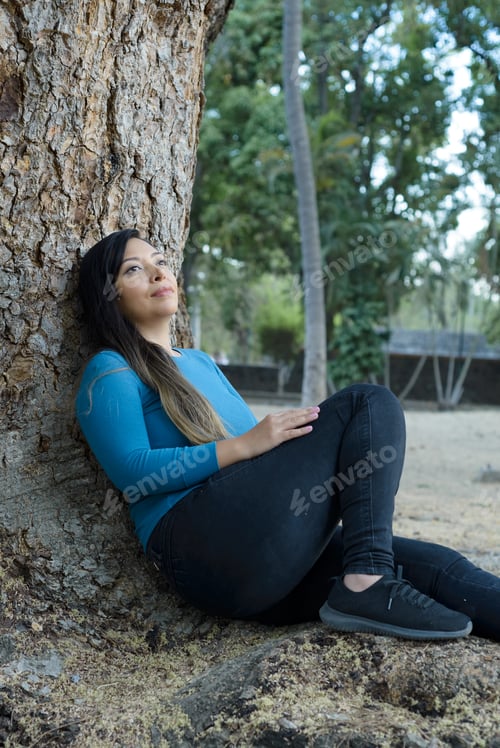 Preview: Woman seated leaning against a tree with pensive attitude.