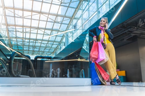 Preview: Mature woman talking smartphone with shopping bags in the mall.