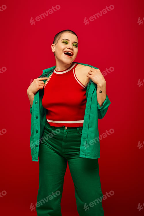Preview: fashion trend, excited and tattooed, short haired woman in green outfit smiling on red background,