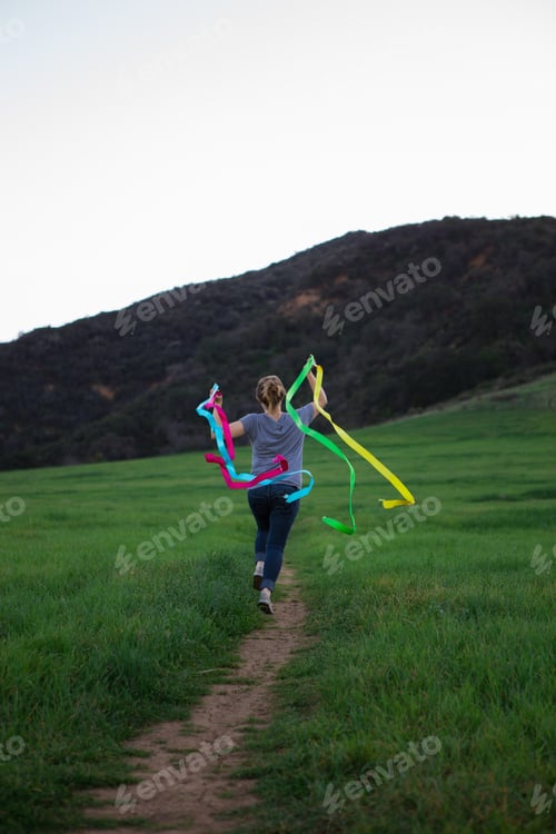 Preview: Rear view of young woman running on path holding up dance ribbons