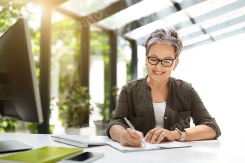 Preview: Positive middle aged woman working at home office, taking notes