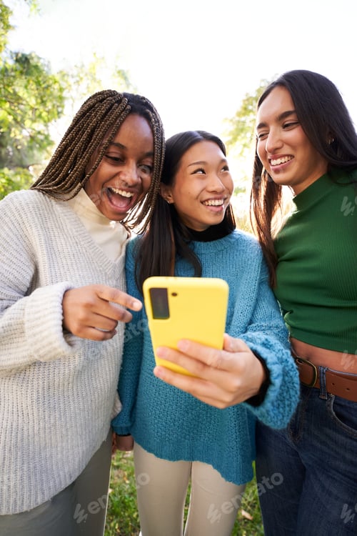 Preview: Vertical. Three young women using phone while having fun in the park and laughing. Social media app