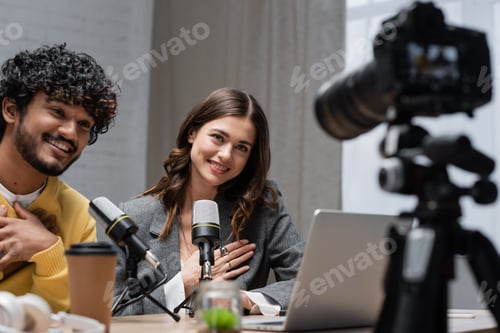 Preview: pleased multiethnic radio hosts looking at blurred digital camera near microphones and takeaway