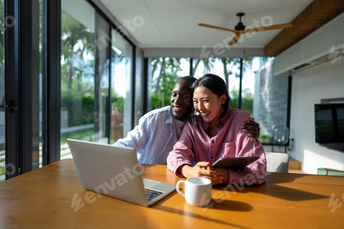 Preview: Young happy multiethnic couple having fun while using laptop, digital device at home