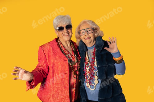 Preview: Two senior women waving and smiling on yellow background