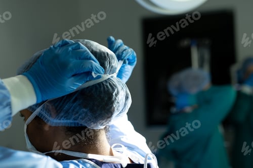 Preview: Rear view of biracial female surgeon in gloves and cap tying mask in theatre, with copy space