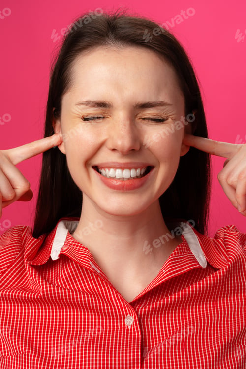 Preview: Woman smiles while covering her ears with fingers against a pink background