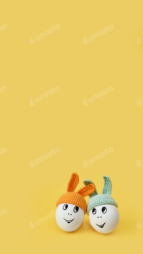 Preview: two white eggs in beanie with rabbit ears and cheerful faces on yellow background