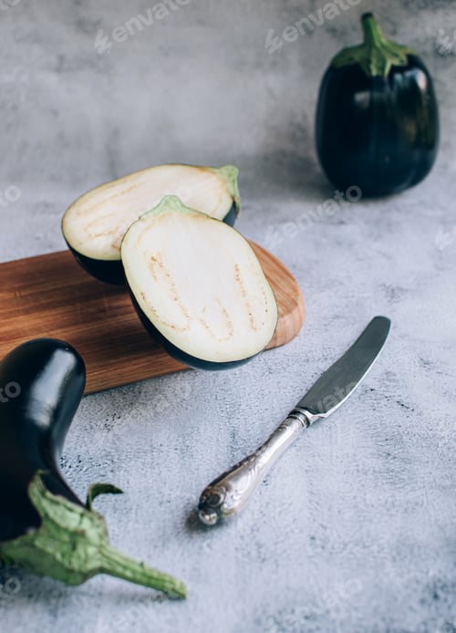 Preview: Halved eggplan on chopping wooden board and knife over gray background.