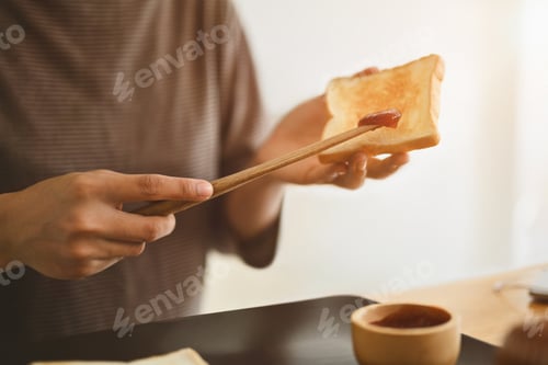Preview: Young man preparing a healthy breakfast spreading strawberry jam on slice of bread