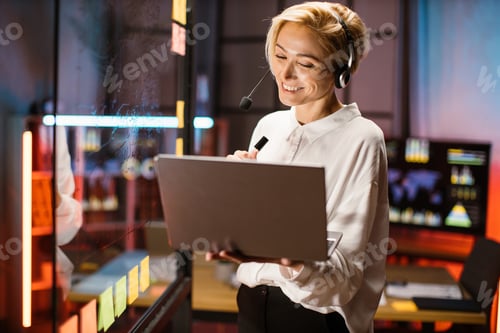 Preview: Businesswoman working at office near glass wall, using headset and laptop