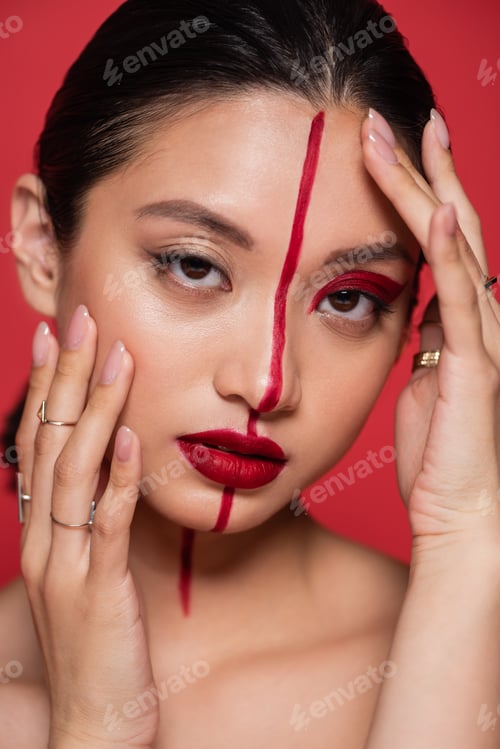 Preview: portrait of brunette asian woman looking at camera while touching face with creative visage isolated