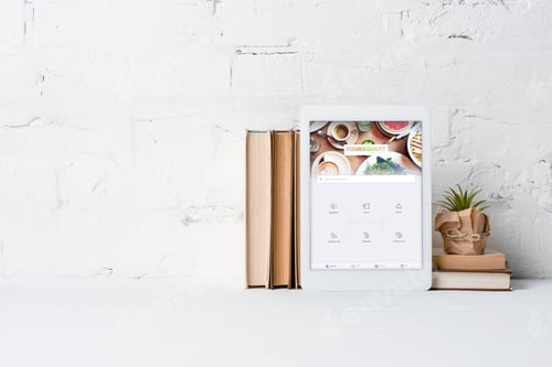 Preview: digital tablet with foursquare application, books and potted plant near white brick wall
