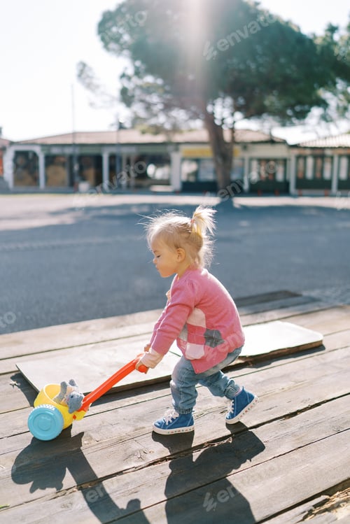 Preview: Little girl is pushing a toy cart with a soft toy on a wooden deck in the park