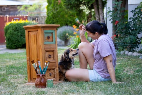 Preview: Woman painting a wooden furniture while sitting with her dog in the garden. DIY concept.