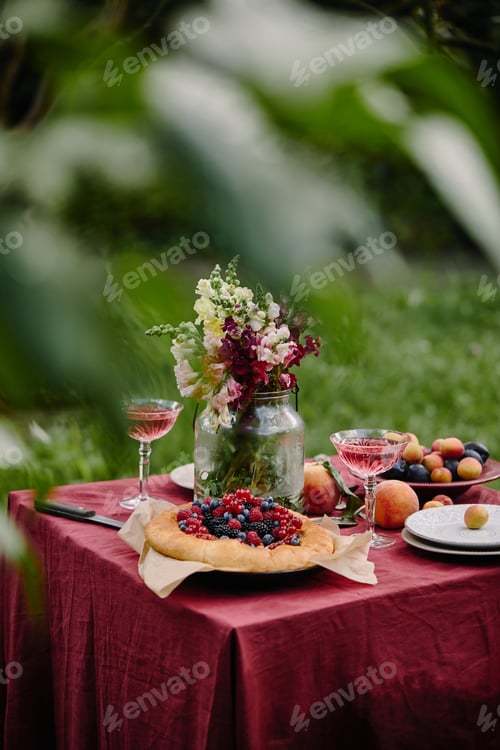 Preview: bouquet of flowers in glass jar, berries pie and wineglasses on table in garden