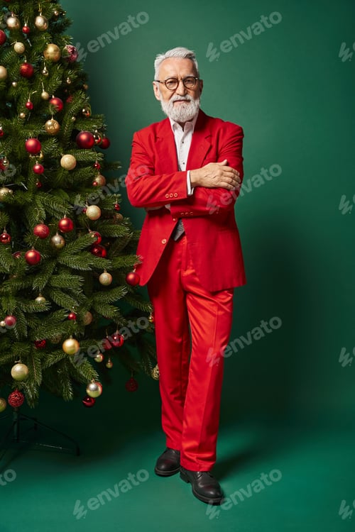 Preview: handsome Santa with white beard and glasses posing near fir tree with crossed arms, winter concept