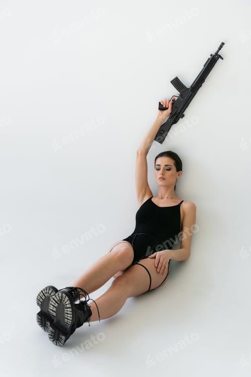 Preview: sexy woman in black bodysuit and boots holding rifle in raised hand while sitting on grey