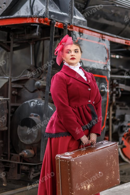 Preview: A girl in a burgundy suit and a hat with a veil stands with a suitcase near an old steam locomotive