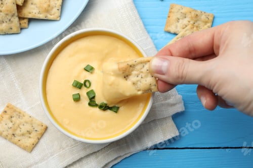 Preview: Woman dipping cracker into tasty cheese sauce at light blue wooden table, top view
