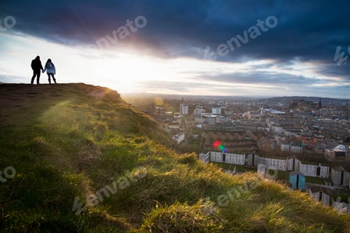 Preview: A young couple hold hands in front of the view of the City of Edinburgh from Salisbury Crags