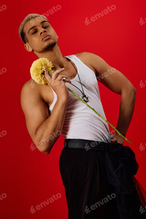 Preview: Trendy young man in tank top showcases style with flower in vibrant red studio