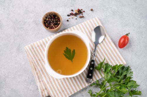 Preview: Meat broth with parsley in a white plate on a light background with fresh herb, spoon and napkin.