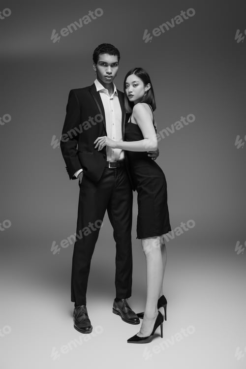 Preview: full length of african american man in elegant suit and asian woman in black dress looking at camera