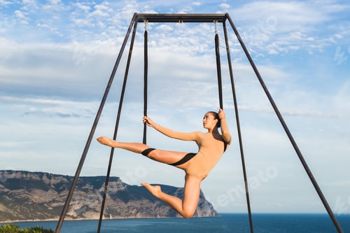 Preview: Woman practicing fly-dance yoga poses in a hammock on the coast with views of the mountains