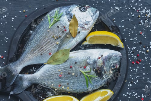 Preview: flat lay with raw fish with lemon, bay leaves and rosemary in tray on black table covered by salt