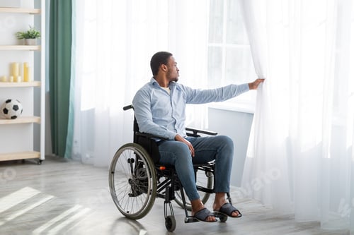 Preview: Sad disabled young man sitting on wheelchair alone at home, looking through window, full length