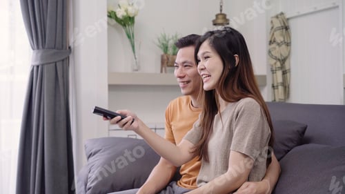 Preview: Asian couple watching TV and drinking warm cup of coffee in living room at home.