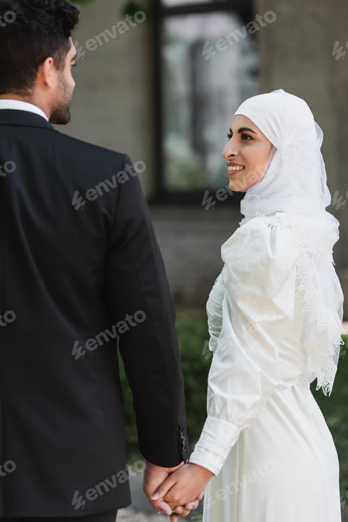 Preview: groom holding hands with smiling muslim bride in wedding dress outside