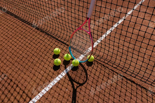 Preview: Tennis racket and balls on clay court near net shadow in sunlight