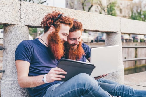 Preview: Young male hipster twins with red hair and beards sitting on bridge browsing digital tablet