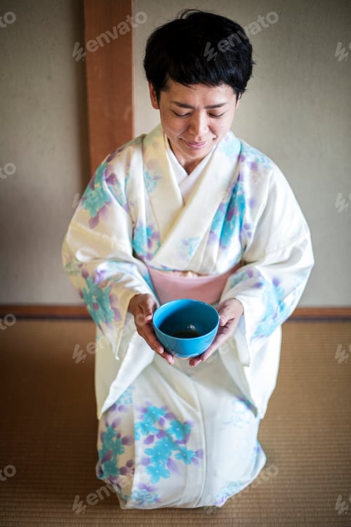 Preview: Japanese woman kneeling on floor during tea ceremony, holding blue tea bowl.