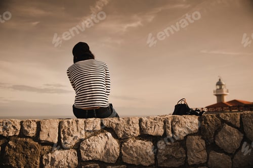 Preview: Back view of elegant lady in cap sitting on meadow near beacon on shore and wonderful sky
