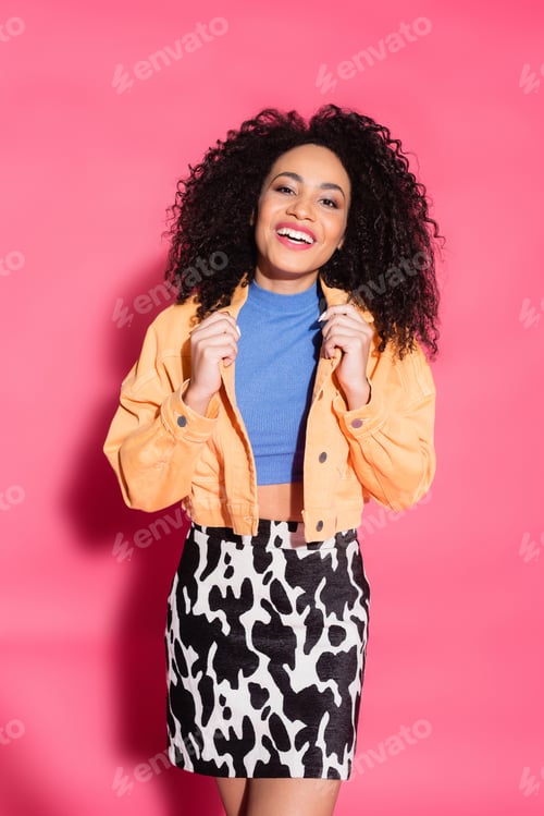 Preview: happy african american woman in skirt with animal print adjusting jacket while posing on pink