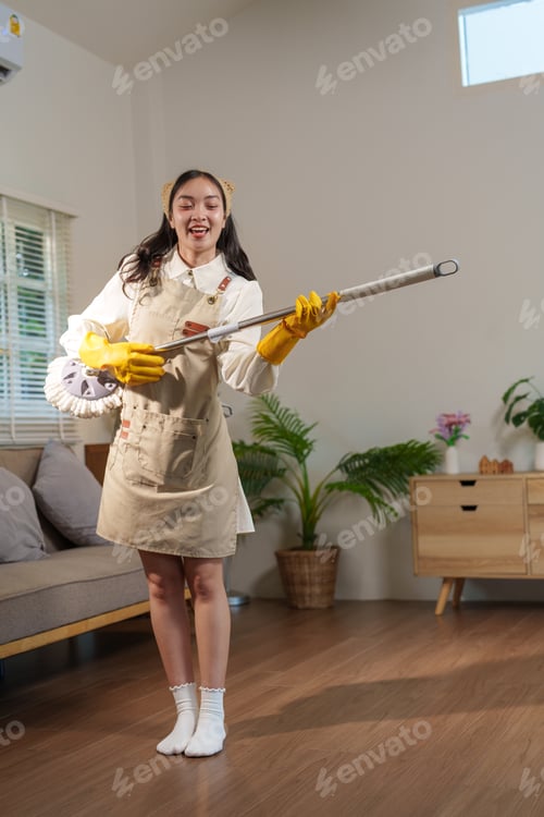 Preview: Young asian woman wearing apron and gloves holding a mop like playing electric guitar and smiling wh