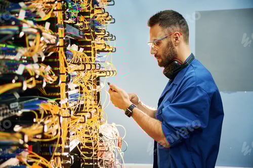 Preview: Side view. Young man is working with internet equipment and wires in server room