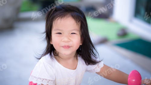 Preview: Adorable Asain child girl is sweet smiling. Kid playing electronic motorcycle toy in front of house.